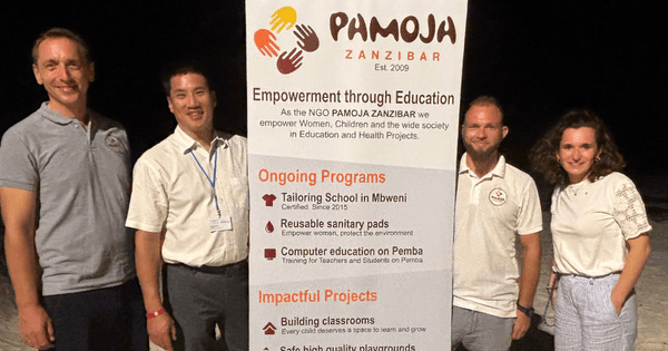 Helen Schröder und Michael Sawang stehen am Abend auf Sansibar gemeinsam mit zwei NGO-Vertretern neben einem Banner der Organisation Pamoja Zanzibar, einem von HOPE geförderten Projekt. Das Foto zeigt die Gruppe am Strand und verweist auf Bildungs- und Gesundheitsprojekte für Frauen, Kinder und die lokale Gemeinschaft.