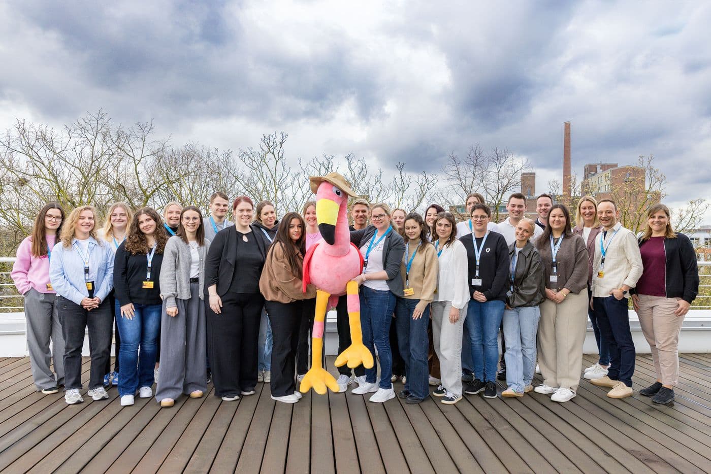 Gruppenfoto beim Azubi-Workshop: Die Teilnehmenden und Betreuenden stehen auf der Dachterrasse von schauinsland-reisen in Duisburg eng beieinander und posieren gemeinsam mit einem pinken Flamingo-Maskottchen vor bewölktem Himmel.