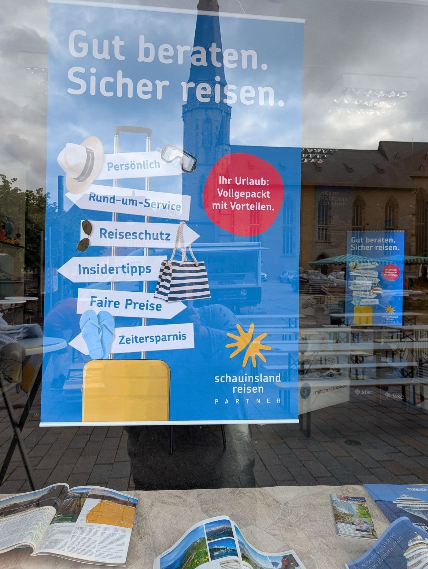 Ein Plakat, das die Reisebürostärken aufzählt hängt in einem Schaufenster. Im Schaufenster spiegelt sich eine Kirche.