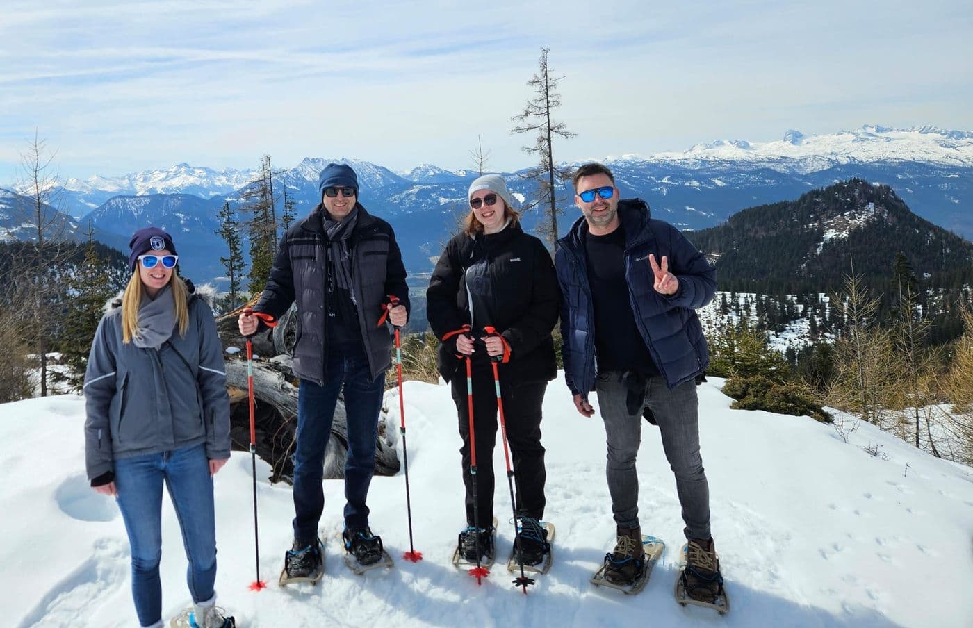 Vier Teilnehmende beim Schneeschuhwandern in Österreich stehen auf einem verschneiten Aussichtspunkt vor einer beeindruckenden Alpenkulisse und posieren mit Stöcken in die Kamera.